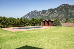 A swimming pool with a wooden structure in the background, surrounded by a grassy lawn and a hedge.