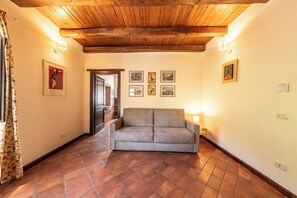 A room with a wooden ceiling, a grey sofa, tiled flooring, and framed pictures on the wall. - Dimora Santē – Historic Villa Surrounded by Olive Trees (Campania)