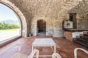 A stone-walled interior with a view of mountains, a table, chairs, and a staircase.