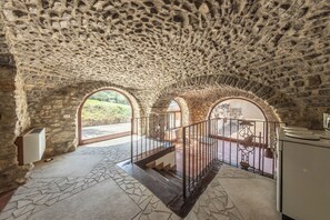 A stone-walled room with a staircase leading to an upper level, featuring arched windows and a stone floor.