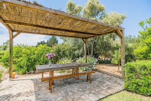 A wooden pergola with a stone bench underneath, surrounded by a garden with various plants and trees. - Rustic Country Retreat for 4 (Pollença)