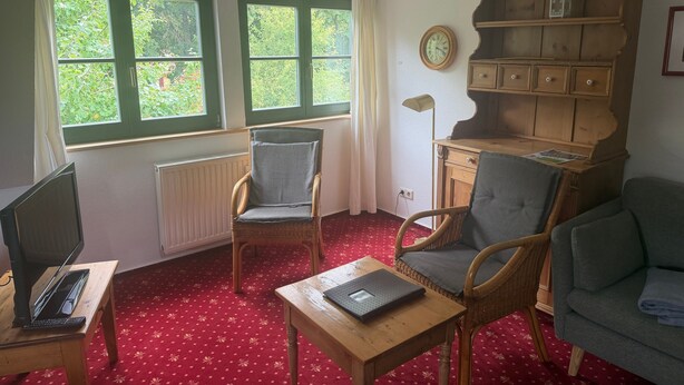 A room with two windows, a wooden bookshelf, a desk with a laptop, two wicker chairs, and a red patterned carpet. - Cozy Holiday Apartment with Garden in Solzow Farmhouse (Südmüritz)