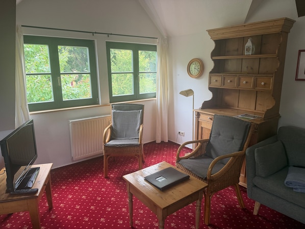 Une pièce avec deux fenêtres, une bibliothèque en bois, un bureau avec un ordinateur portable, deux fauteuils en osier et un tapis rouge à motifs.