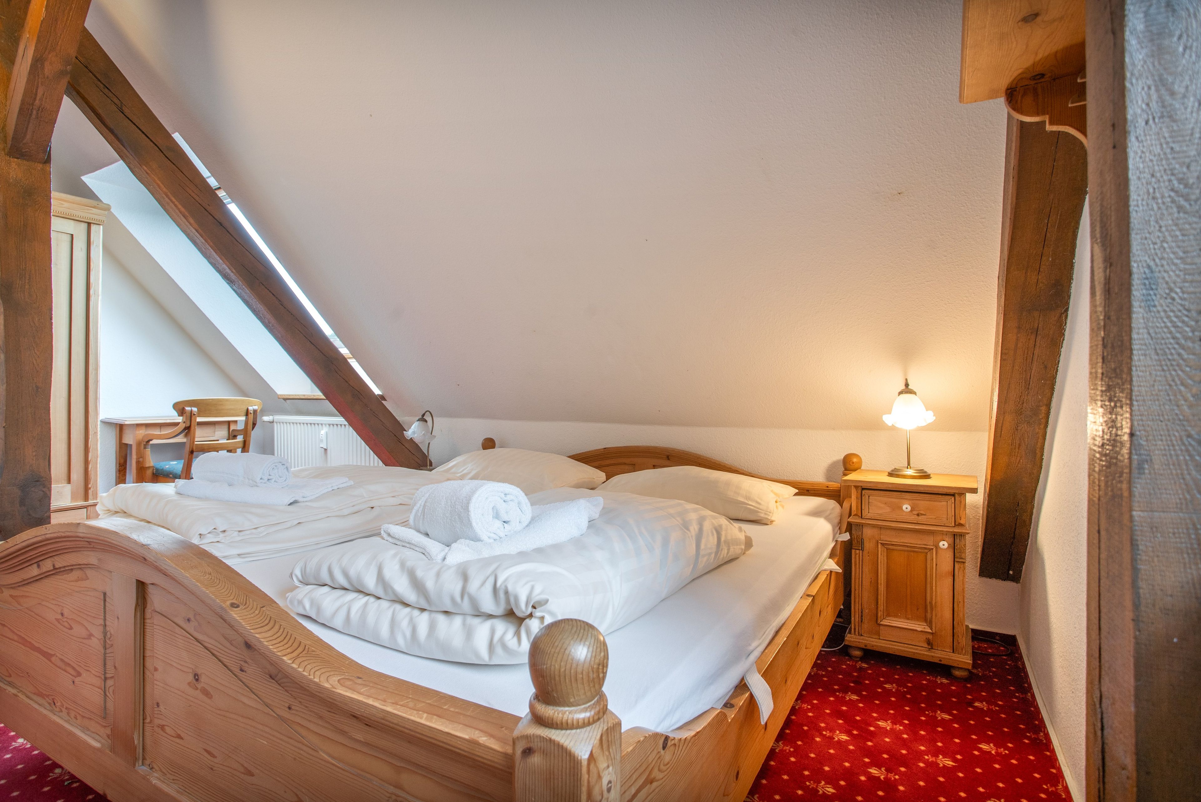 Une chambre à coucher avec un lit en bois, du linge de lit blanc, une moquette rouge, une table de chevet avec une lampe et une lucarne.