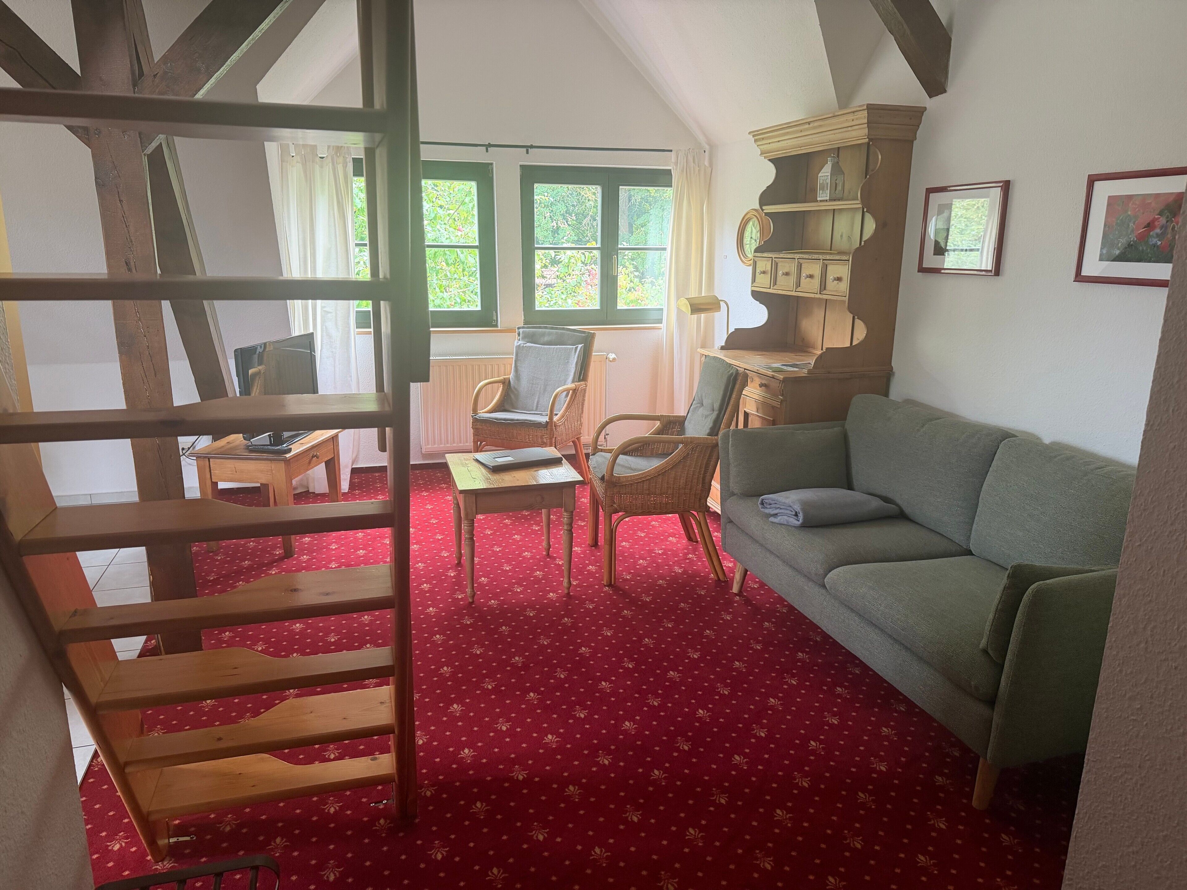 Ein Zimmer mit einer Holztreppe, einem grünen Sofa, einem Korbsessel, einem kleinen Tisch und einem Bücherregal.