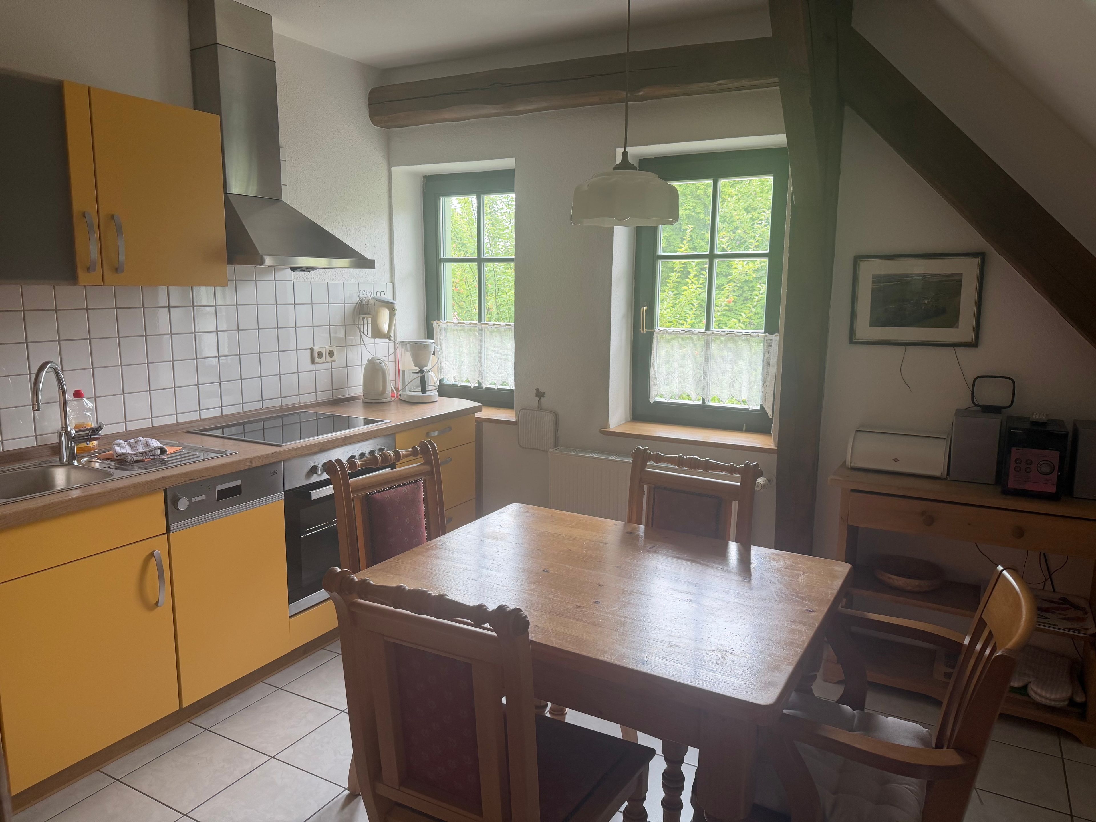 A kitchen with a dining table, chairs, a coffee maker, and a window with a view of greenery.