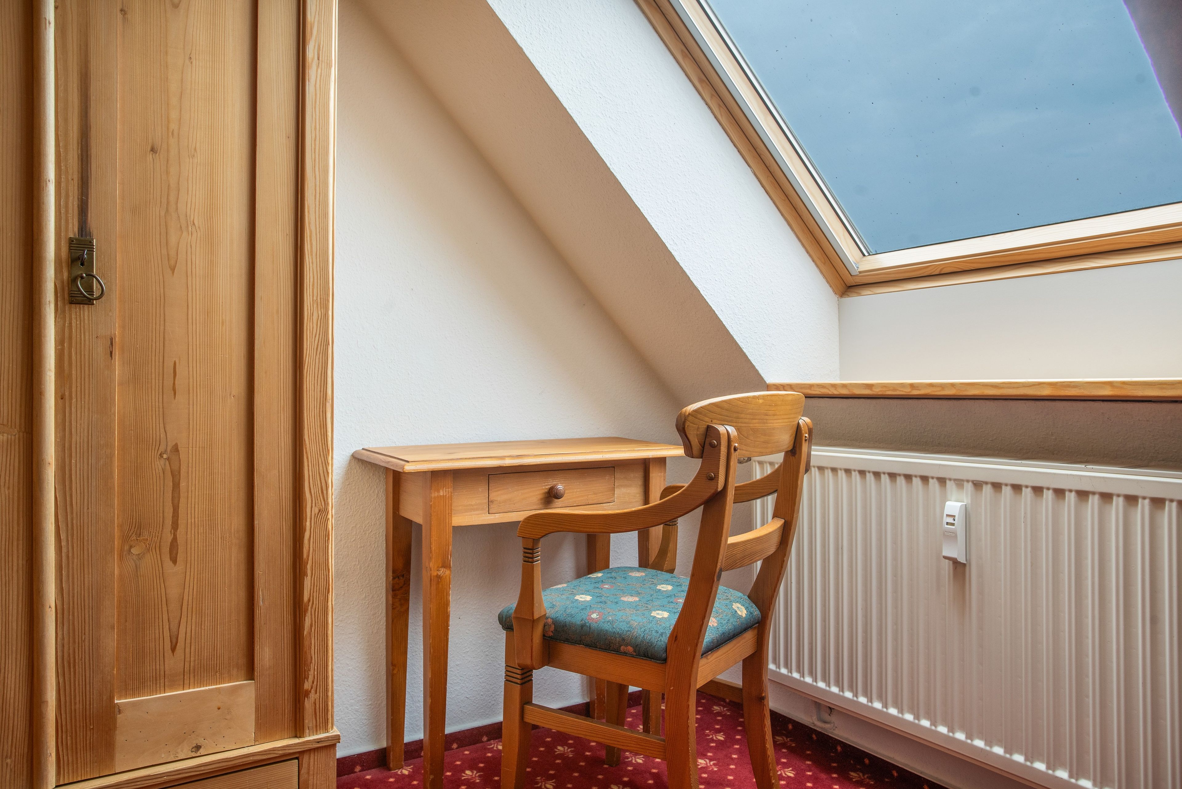 Ein Eckzimmer mit Schreibtisch aus Holz, einem Stuhl mit Blumenkissen, einem Holzschrank und einem Oberlicht.