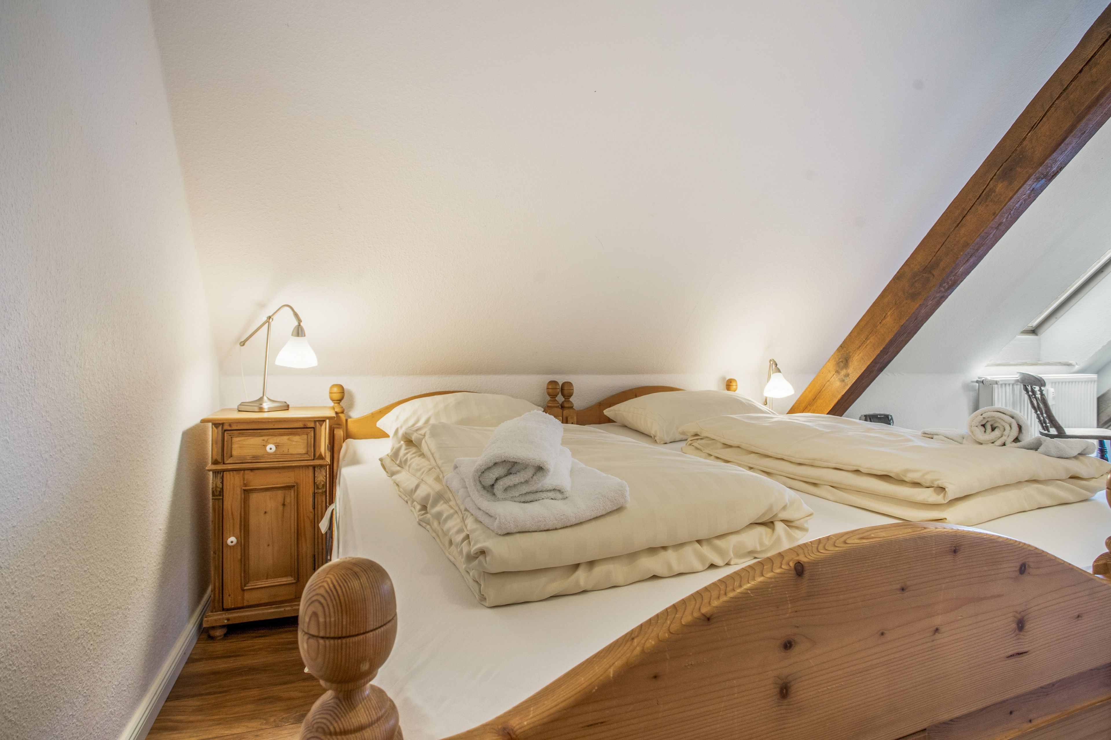 Une chambre avec un plafond en pente, un lit en bois avec deux oreillers, une table de chevet avec une lampe et une serviette enroulée sur le lit.