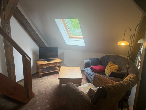 Une pièce confortable avec un téléviseur posé sur un meuble en bois, un canapé avec des coussins, une petite table et une lampe sur pied.