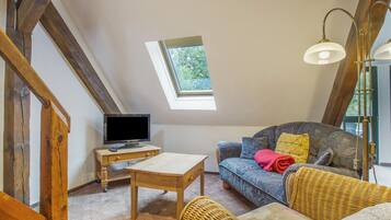 Une pièce chaleureuse avec un plafond en pente, un téléviseur posé sur une table en bois, un canapé agrémenté de coussins à motifs et un fauteuil en osier.