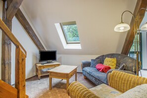 Una habitación acogedora con techo inclinado, un televisor sobre una mesa de madera, un sofá con cojines estampados y una silla de mimbre.