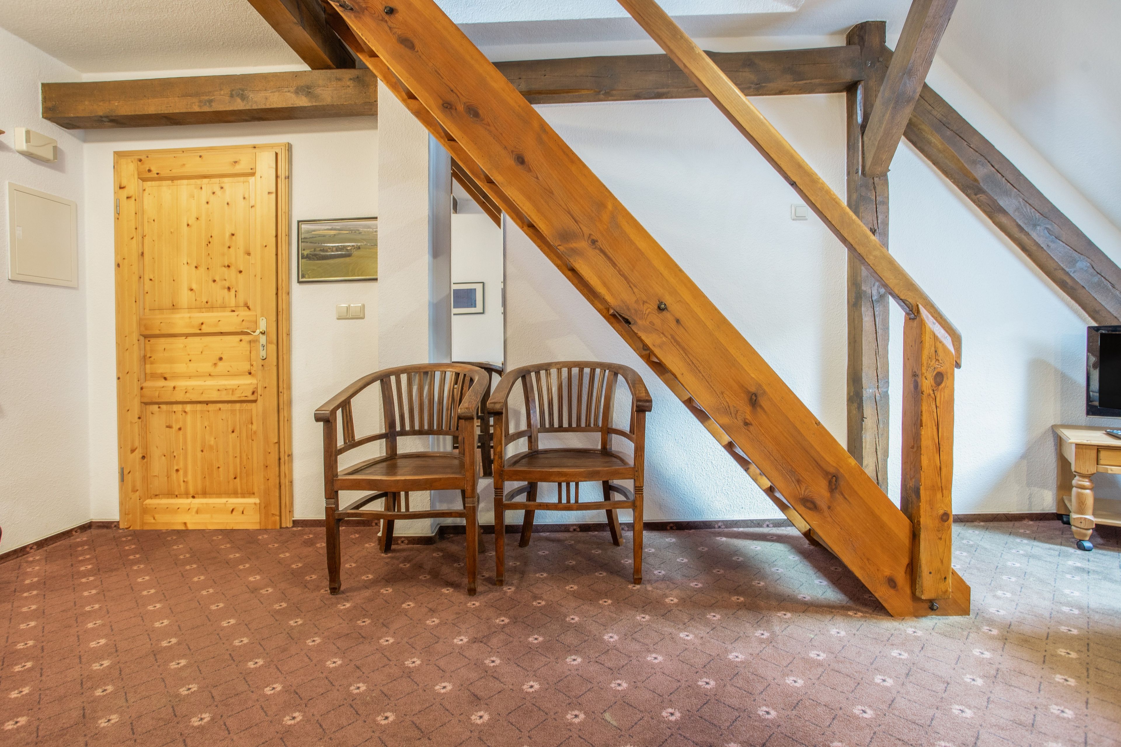 Ein Zimmer mit einer Holztreppe, zwei Holzstühlen, einer Holztür und einem gemusterten Teppich.