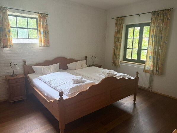 Un dormitorio con cama de madera, ropa de cama blanca, una mesita de noche con lámpara y una ventana con cortinas.