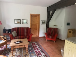Un saló amb un sofà, una butaca, una taula de centre i una catifa. Hi ha quadres amb marc a la paret i una porta de fusta.