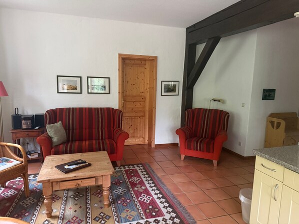 A living room with a sofa, armchair, coffee table, and a rug. There are framed pictures on the wall and a wooden door.
