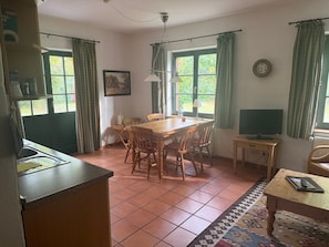 Un coin repas avec une table et des chaises en bois, un téléviseur posé sur un meuble, et un espace cuisine comprenant un évier et des placards.
