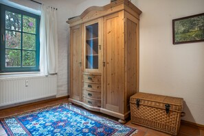 Habitació amb un armari de fusta gran, una finestra amb cortines blanques, una catifes amb dibuixos i un arca de vimet.
