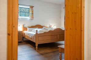 Ein hölzernes Bett in einem Raum mit weißer Backsteinwand, einem Fenster mit floralem Vorhang und einem Holzboden.