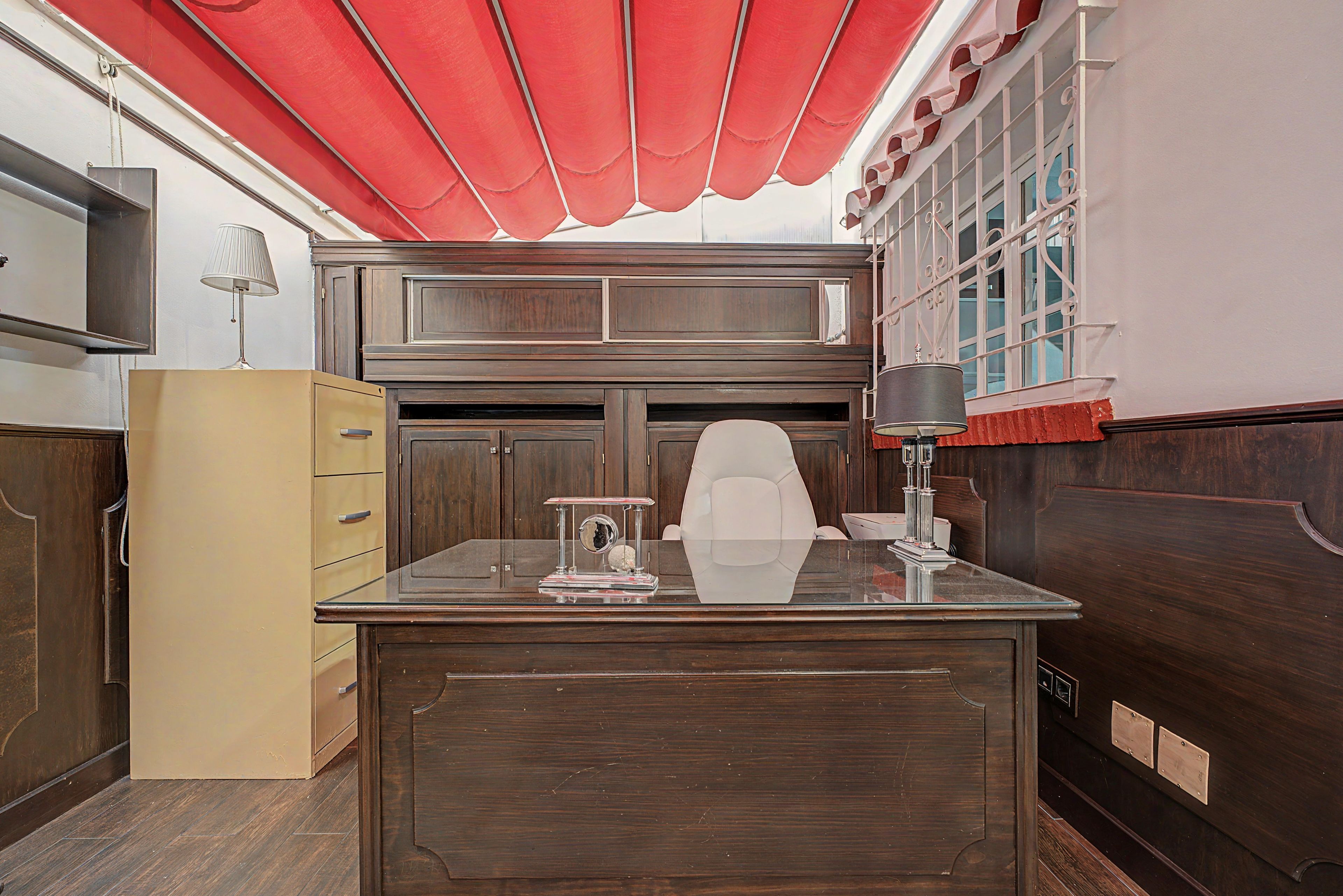 A room with a wooden desk, a lamp, a red awning, and a window with decorative ironwork.