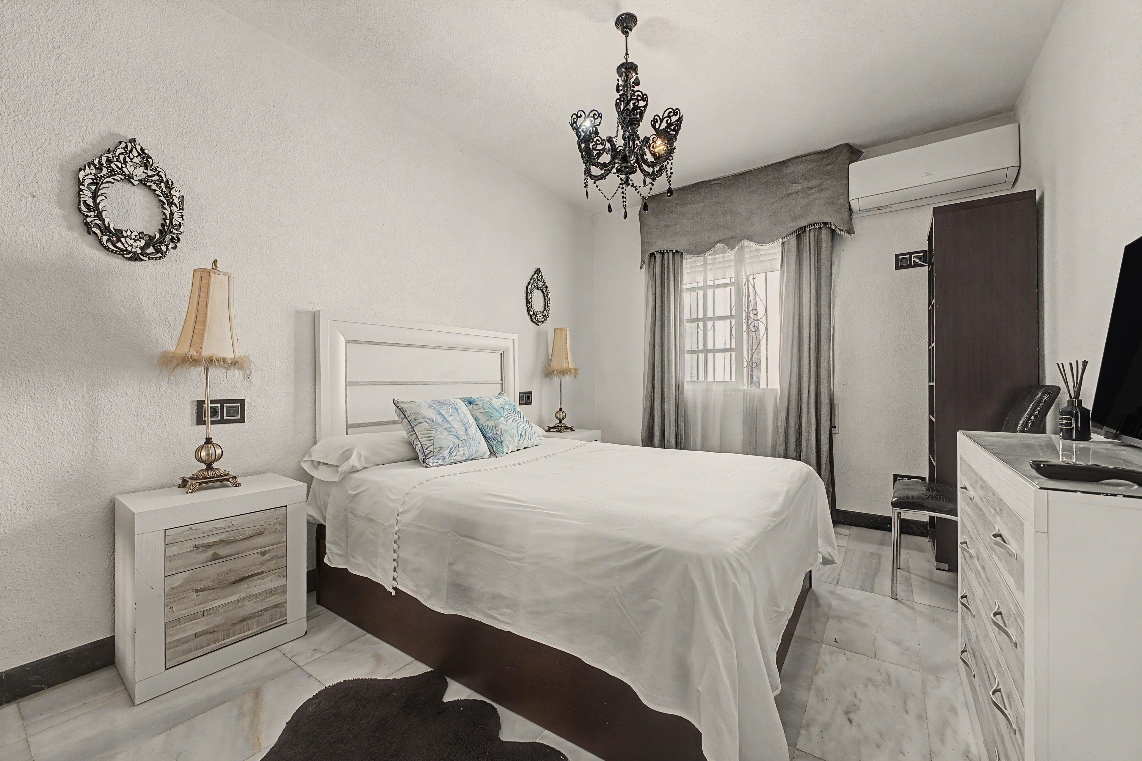 A bedroom with a bed, bedside tables, a dresser, a mirror, a chandelier, and a window with curtains.