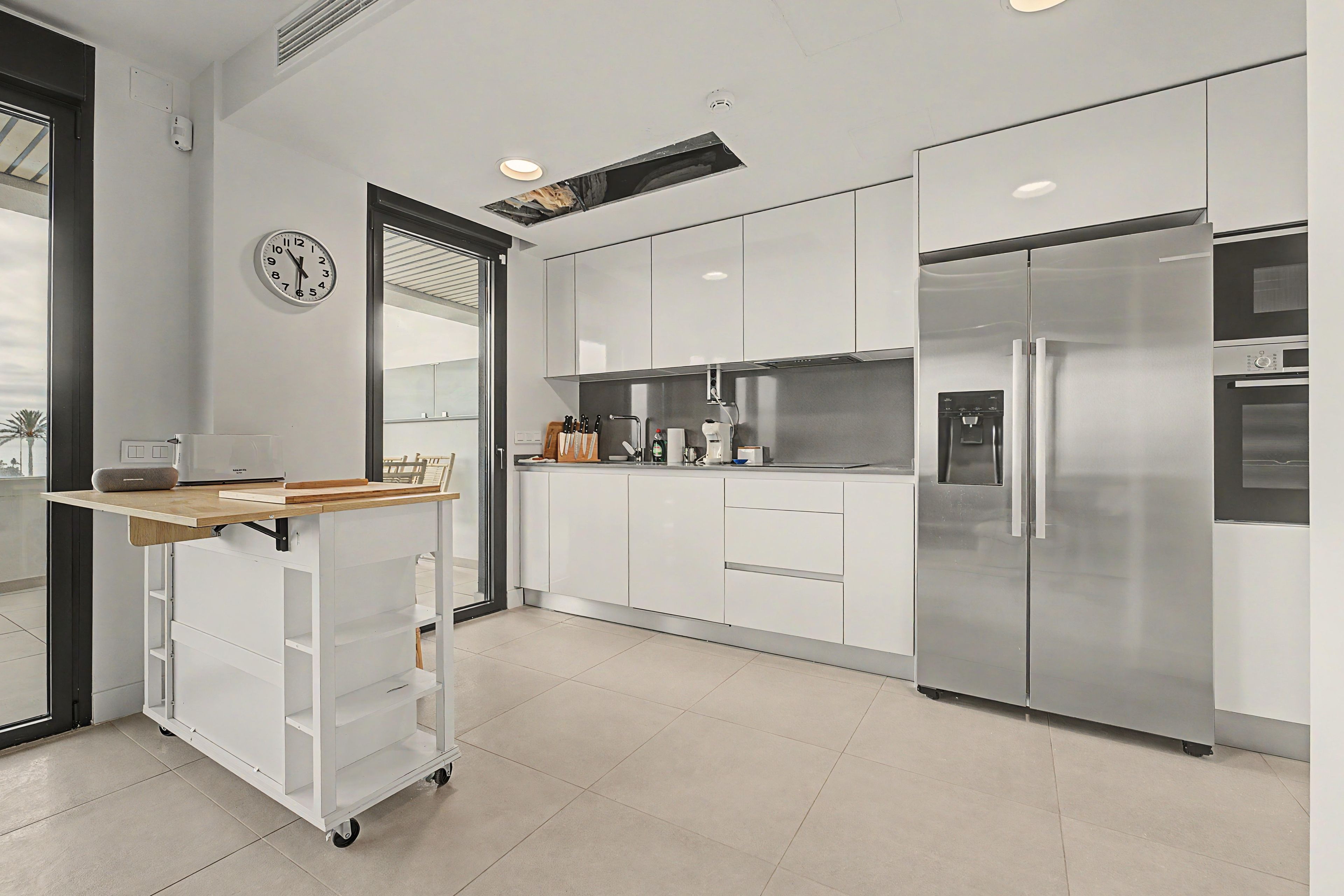Une cuisine moderne avec des meubles blancs, un réfrigérateur en acier inoxydable, une machine à café et une horloge murale.