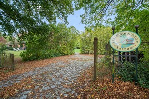 Un chemin pavé de pierres menant à un panneau orné d’un logo et d’un texte, le tout entouré de verdure et d’une clôture.