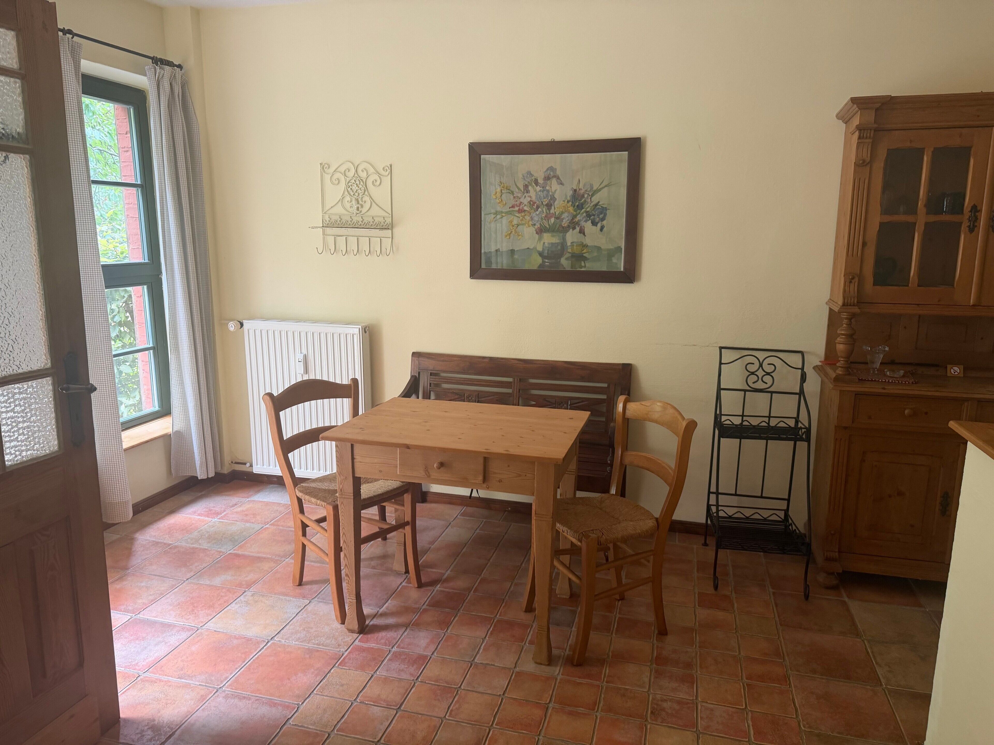 Ein Zimmer mit einem Holztisch, Stühlen, einem Heizkörper, einem Fenster mit Vorhängen, einem Wandbild und einem Holzschrank.