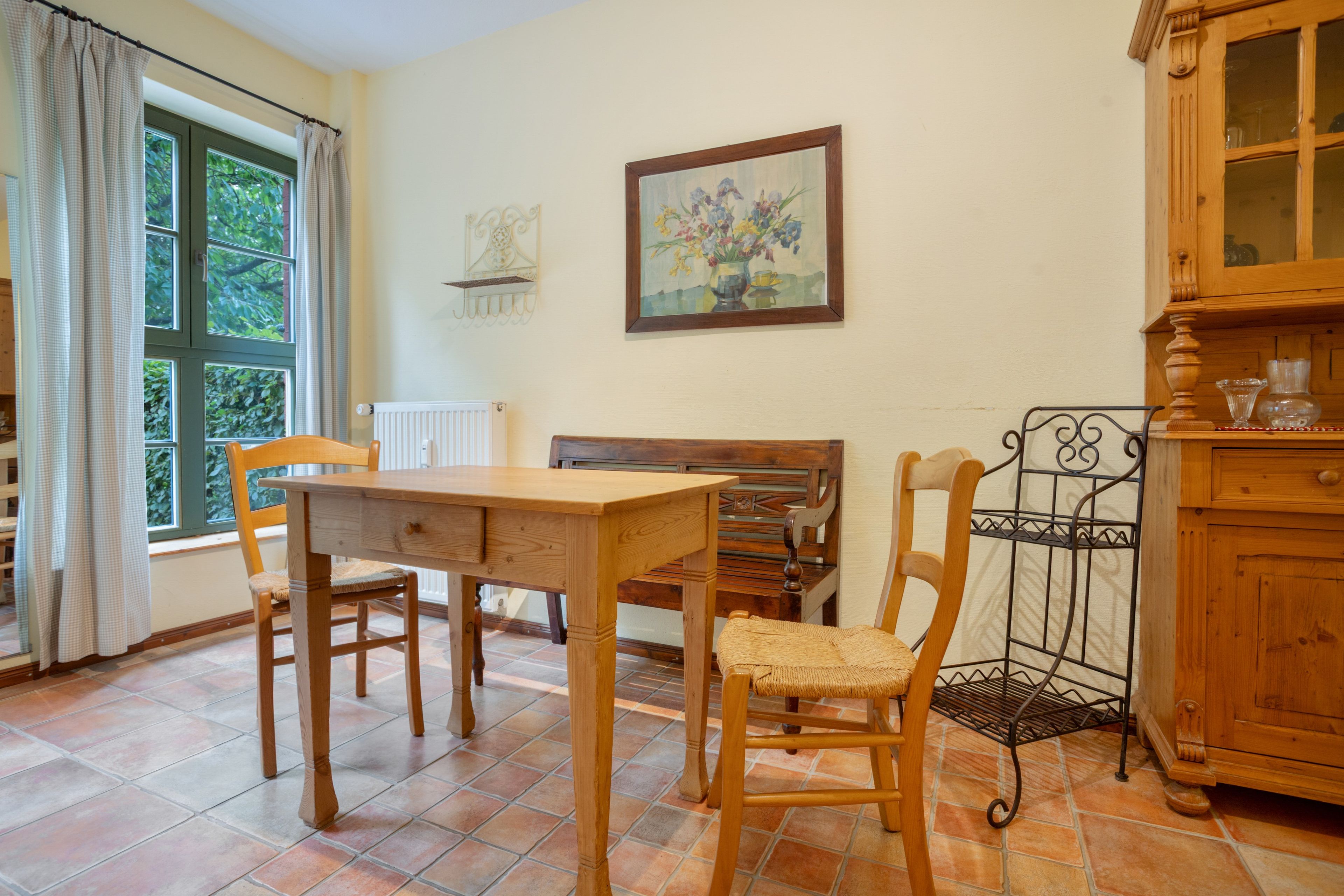 A dining area with a wooden table and chairs, a window with curtains, a radiator, a painting on the wall, and a wooden cabinet.