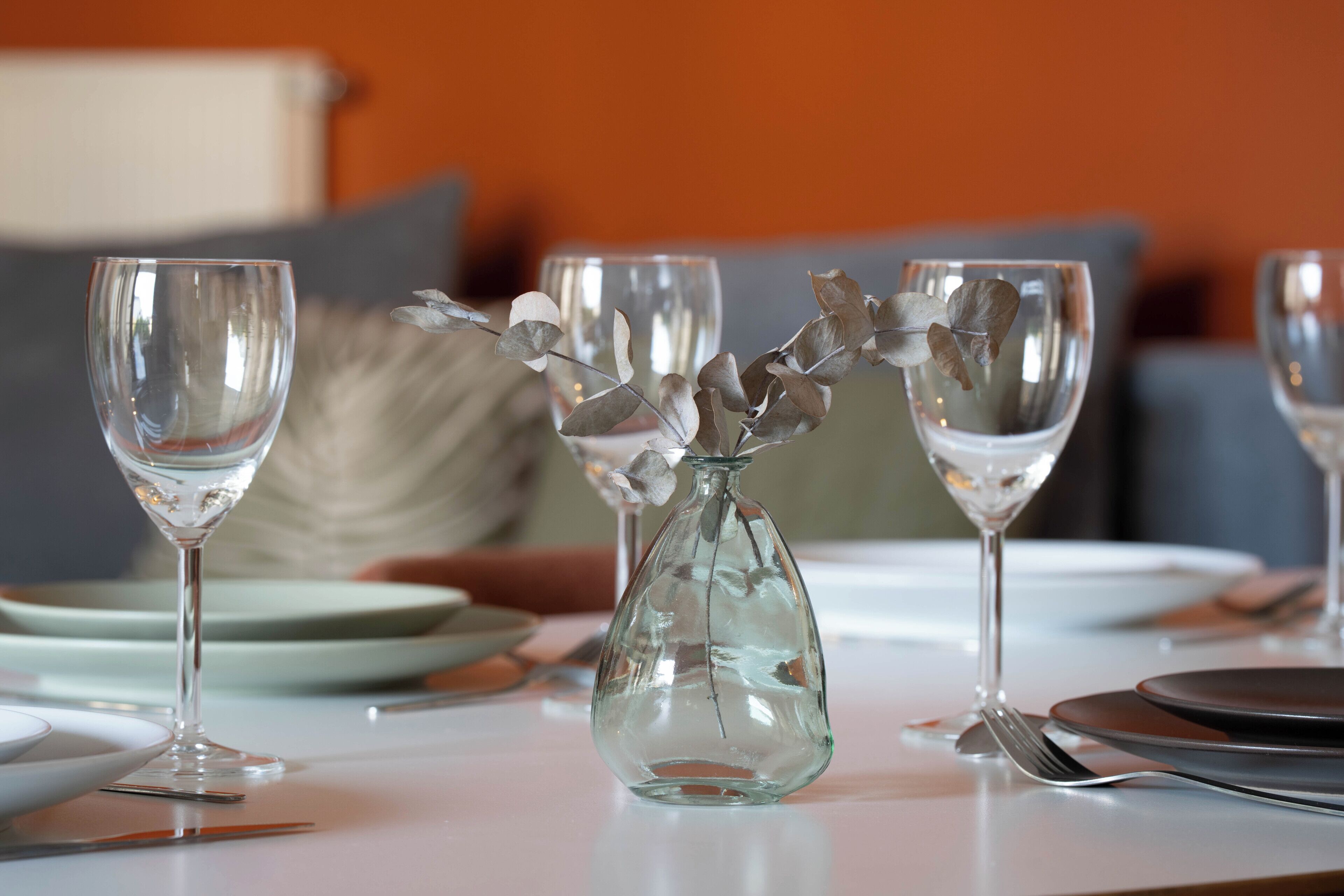 A dining table set with clear glassware and a decorative glass vase with dried leaves.