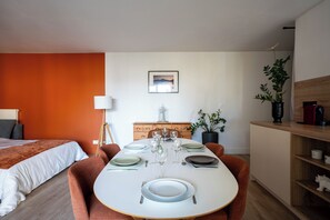 Un espace repas moderne avec une table ronde dressée pour quatre personnes, un meuble en bois, une plante en pot et, au fond, un lit avec une tête de lit blanche.