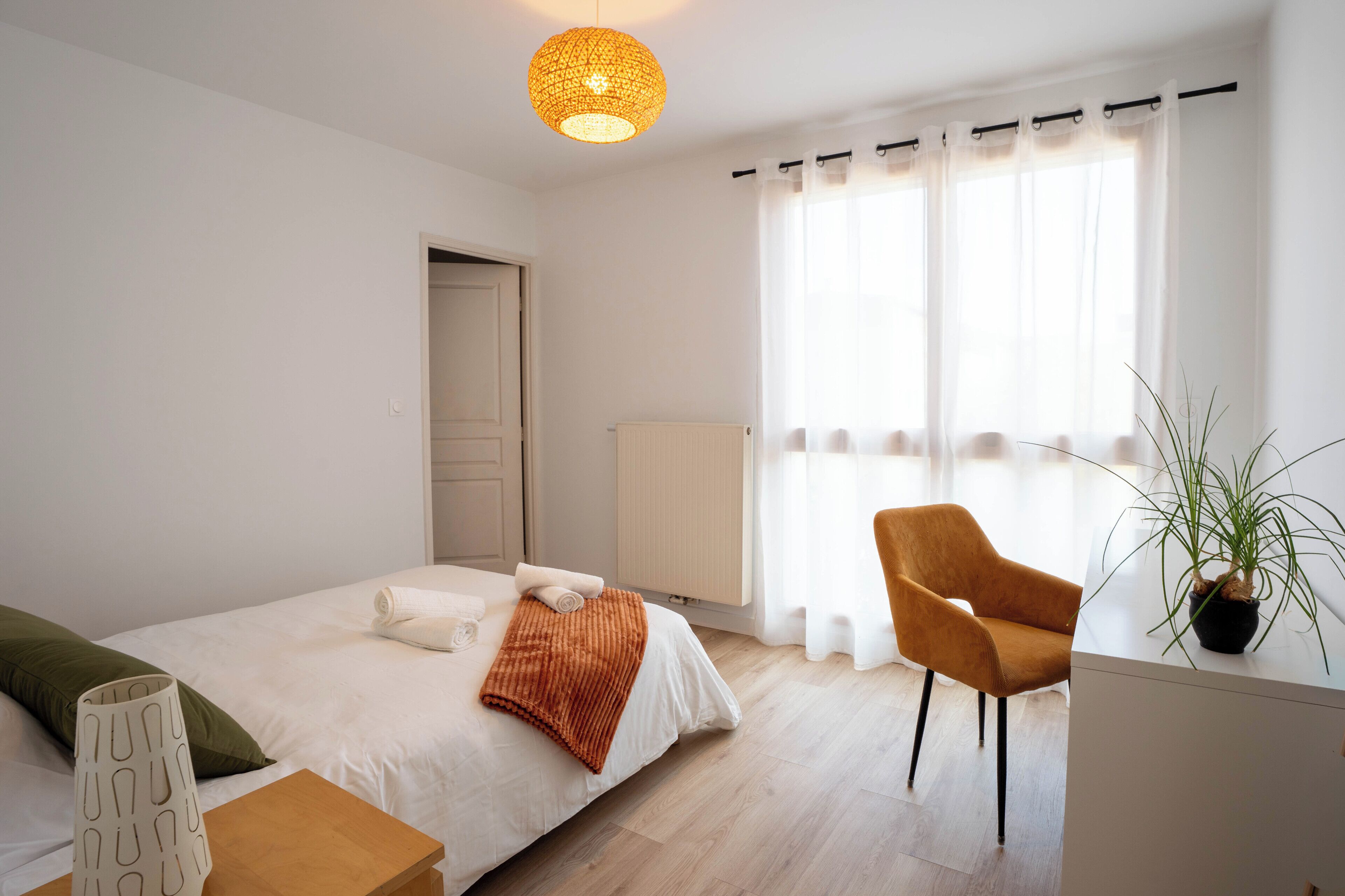 A bedroom with a bed, a chair, a bedside table, a potted plant, and a window with curtains.