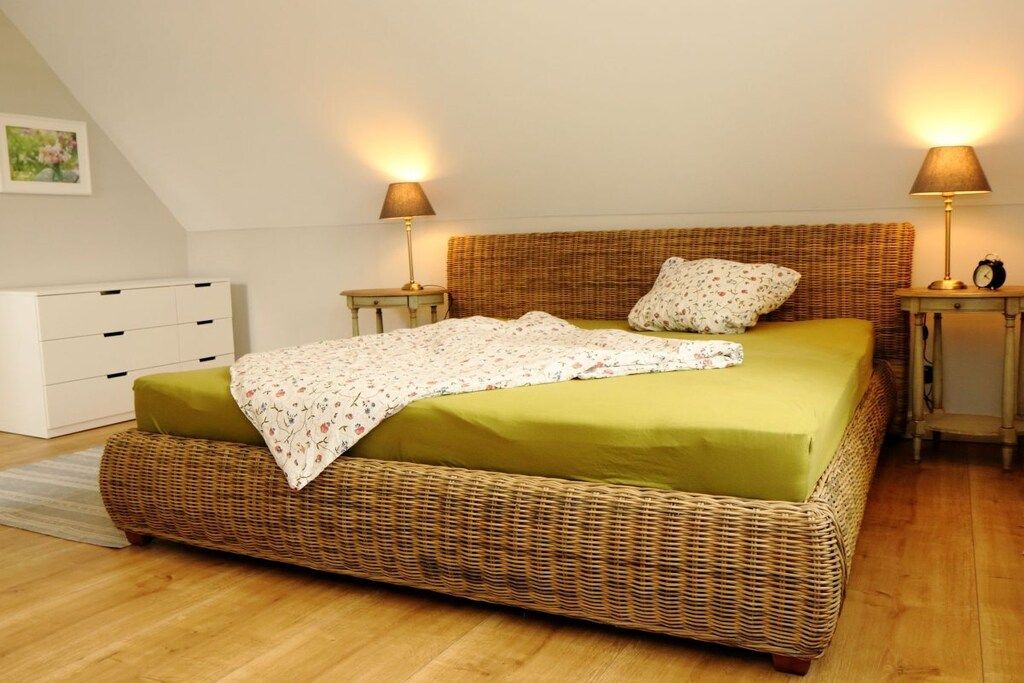 Une chambre à coucher avec un lit en osier, deux tables de chevet avec des lampes, une commode blanche et un tableau encadré au mur.