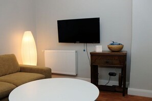 Ein Wohnzimmer mit einer Couch, einem weißen Couchtisch, einem an der Wand montierten Fernseher, einer Heizung und einem Holznachttisch mit einer Schale.