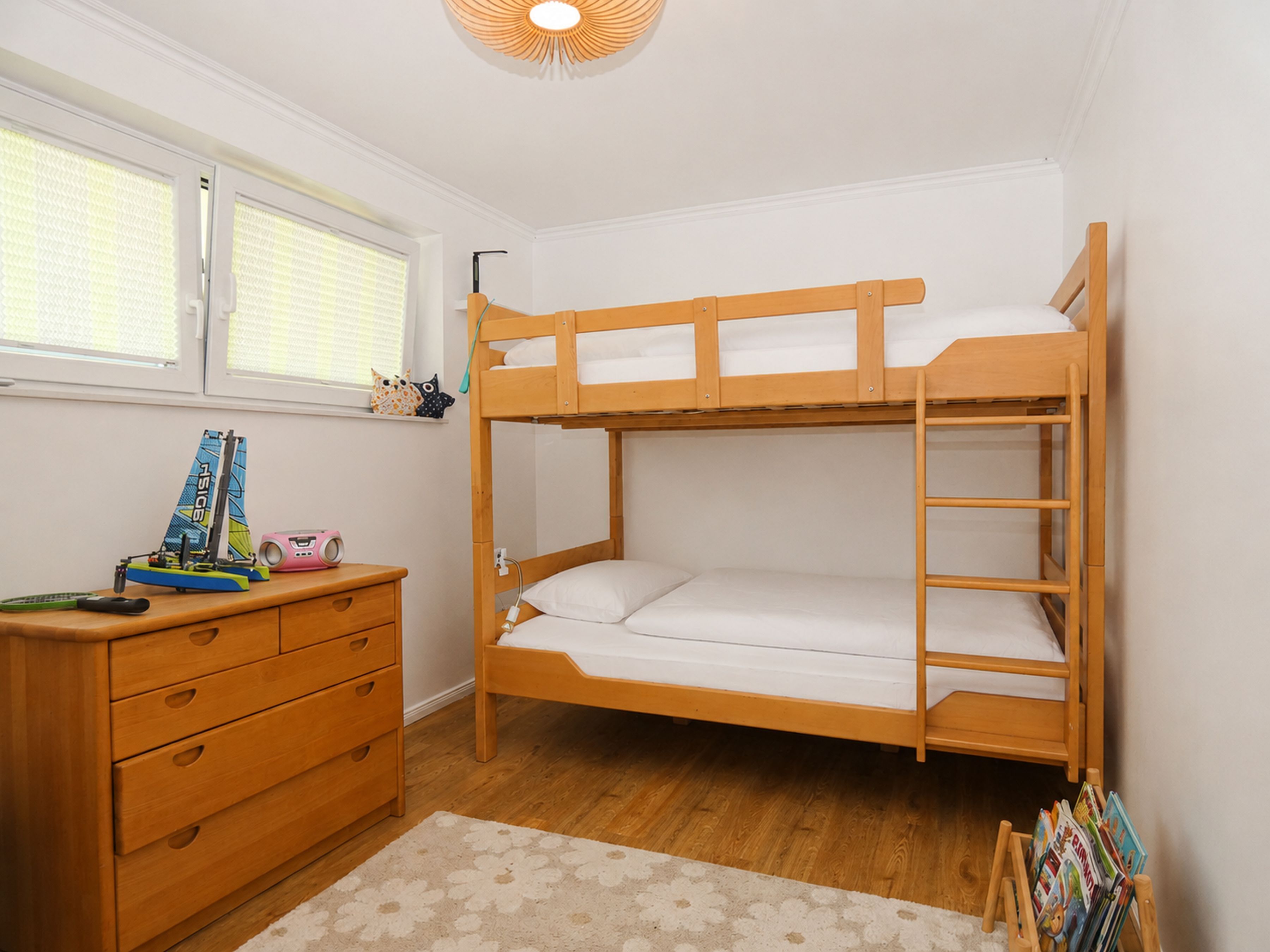 A bunk bed with a wooden frame and white bedding, a wooden dresser with drawers, a window with blinds, and a small shelf with toys and books.