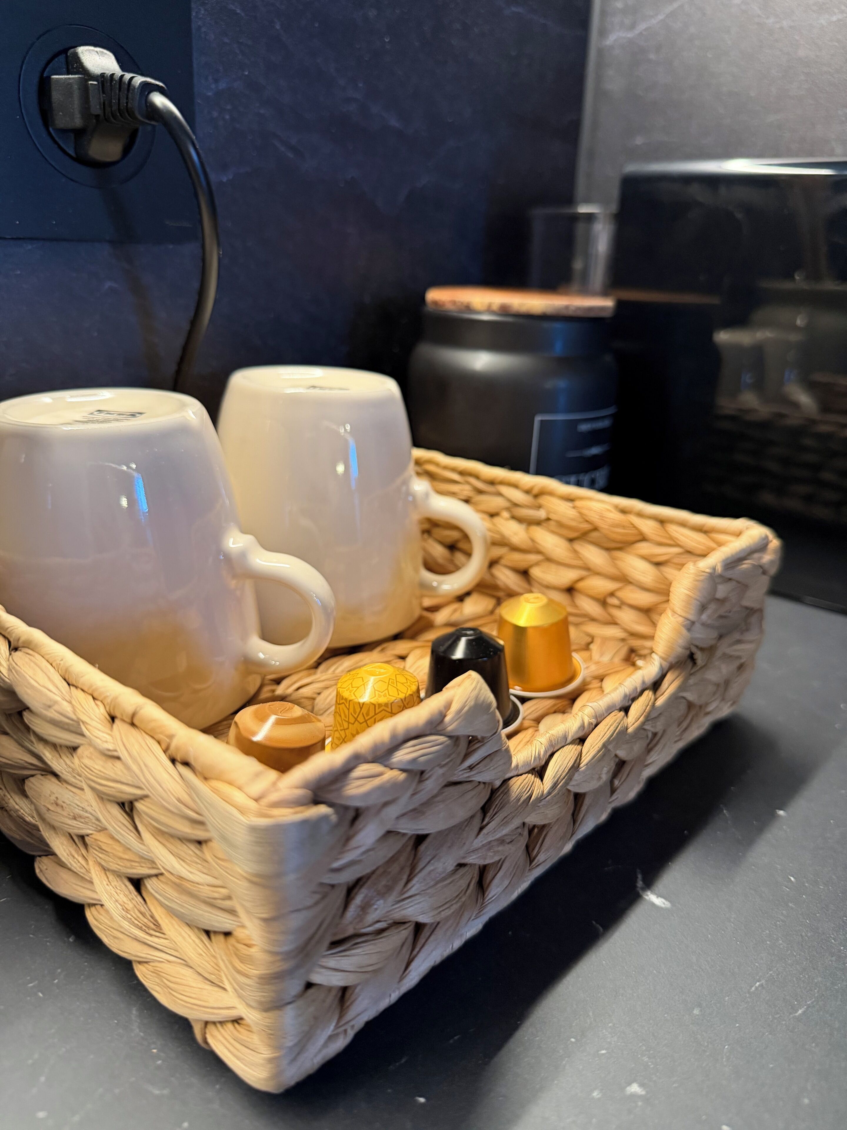 En flettet kurv med to hvide kopper og et sæt te-tilbehør på en bordplade.