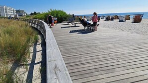 Ein hölzerner Steg führt zu einem Strand, wo Menschen spazieren gehen, ein Rollstuhl zu sehen ist und Strandhütten stehen.