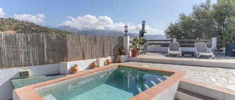 A rectangular pool with a tiled edge, surrounded by a stone patio. There are potted plants and outdoor furniture.