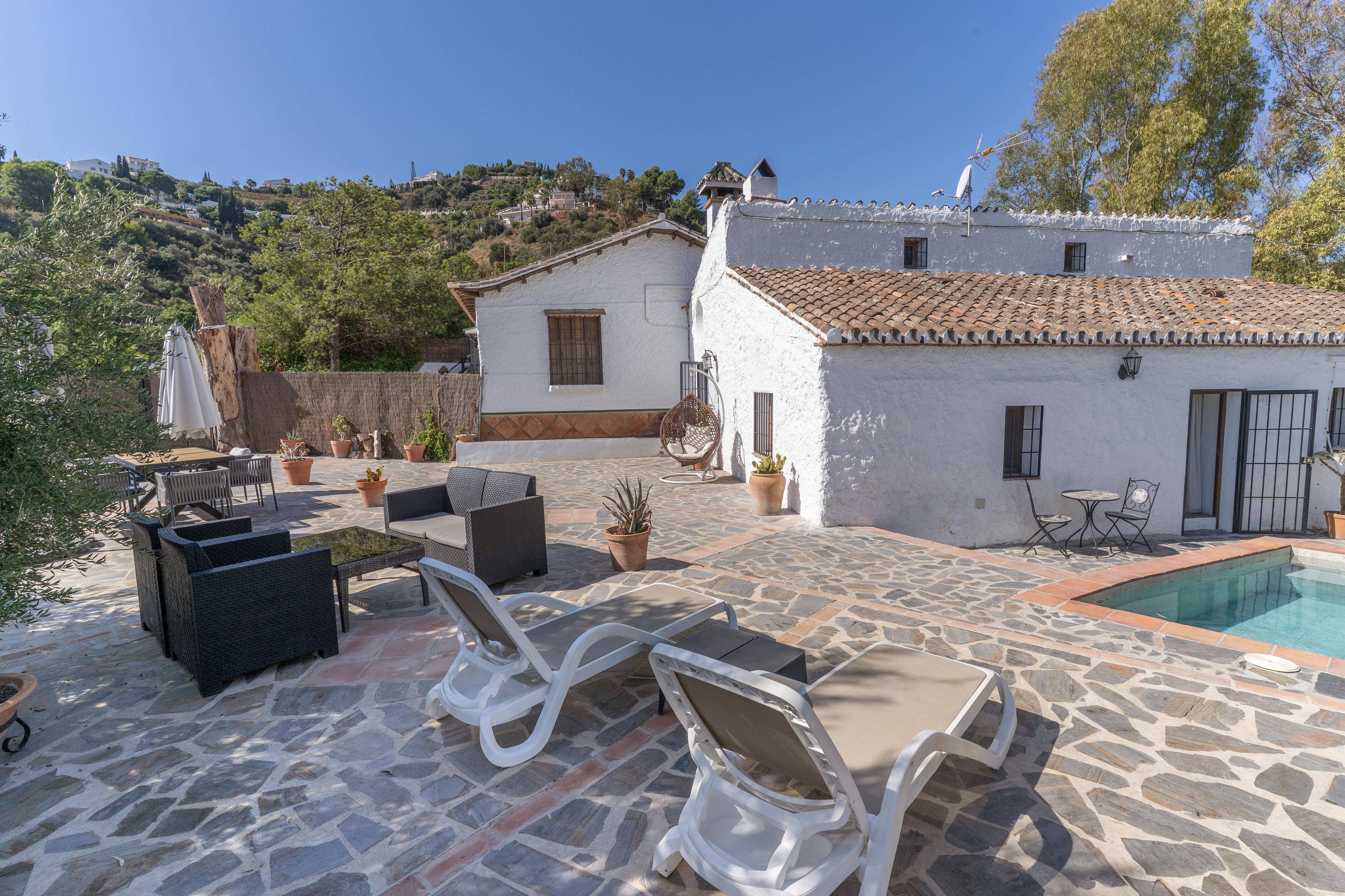 Un patio de piedra con mobiliario exterior, piscina y un edificio blanco con techo de tejas.