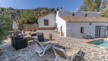 Un patio de piedra con mobiliario exterior, piscina y un edificio blanco con techo de tejas.