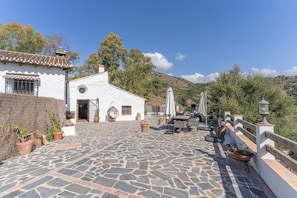 A white building with a tiled roof, a stone patio, and a view of a hilly landscape.