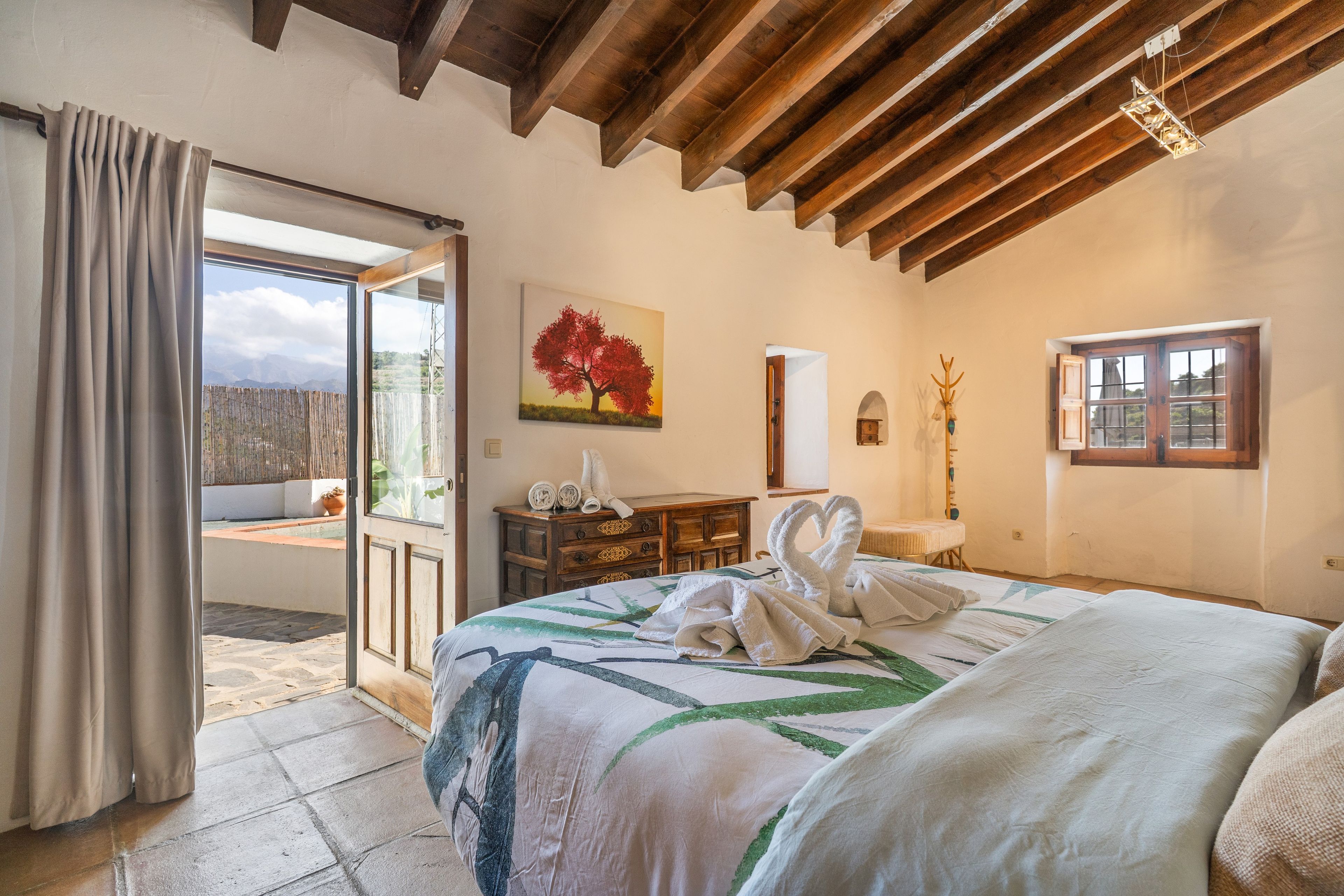 A bedroom with a large bed, a painting, a wooden dresser, and a view of the outdoors.