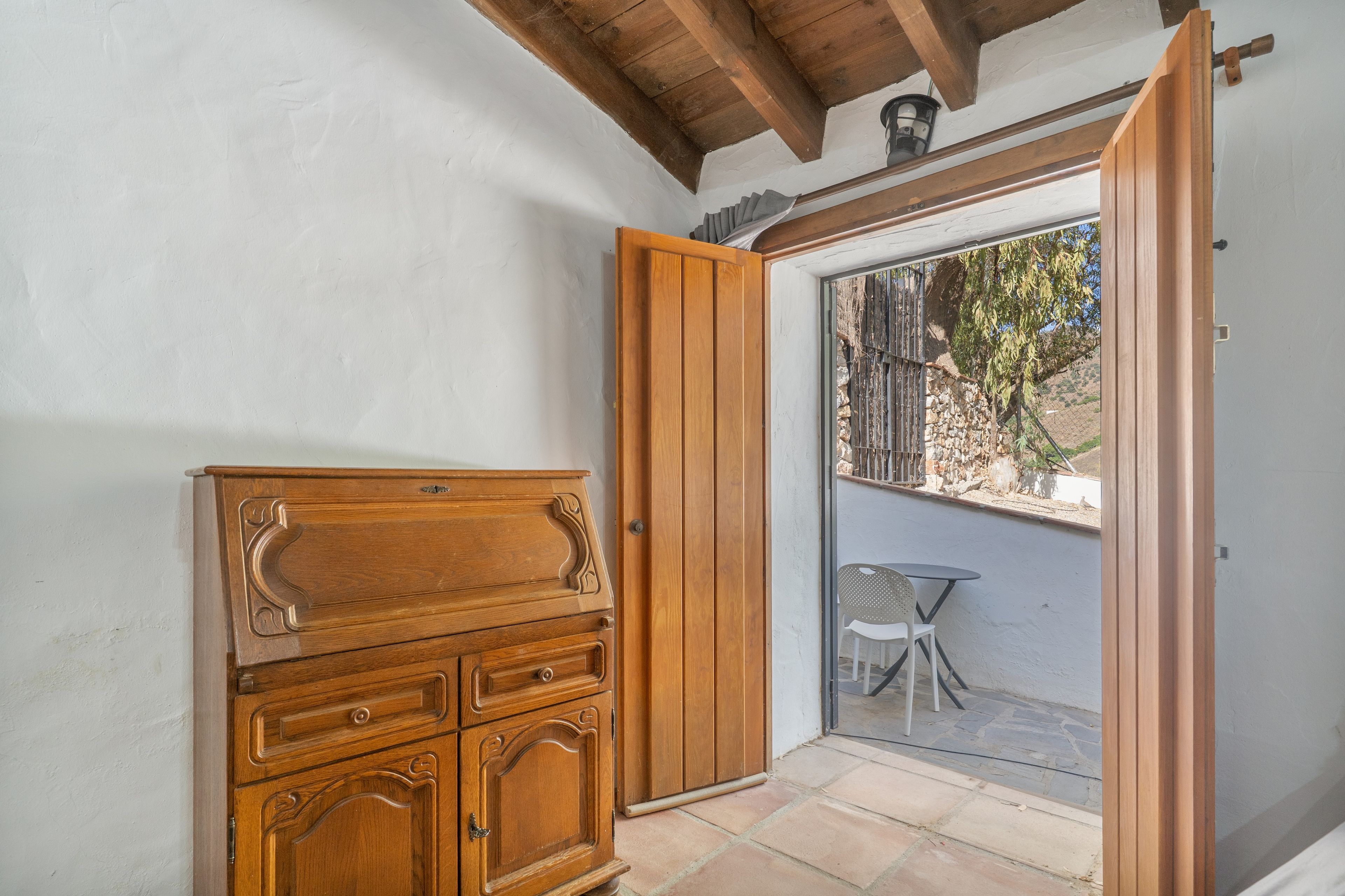 Un mueble de madera con cajones y una puerta, una pequeña mesa con una silla, y una puerta corrediza de vidrio que da a un área exterior con valla y árboles.