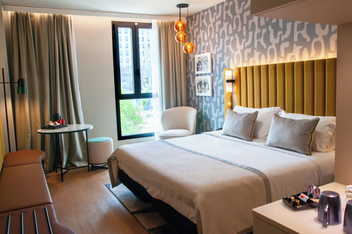 A hotel room with a large bed, a desk, a chair, and a window with a view of buildings.