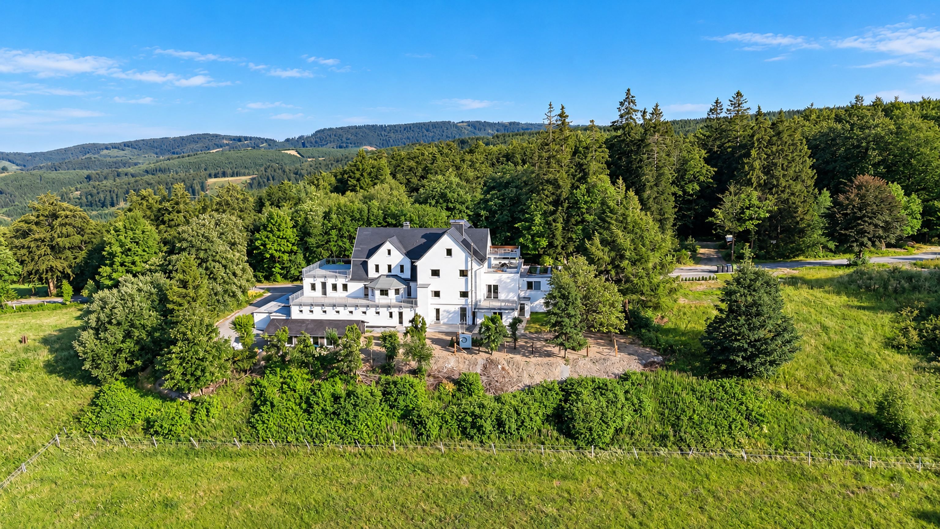 Une grande maison moderne, entourée d’une végétation luxuriante et d’arbres, avec un paysage vallonné en arrière-plan.