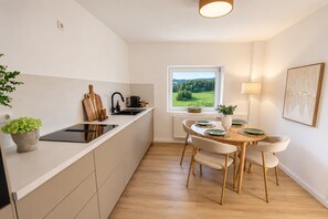 Moderni keittiö, jossa on sisäänrakennettu liesi, ruokailutila pöytineen ja tuoleineen sekä ikkuna, josta avautuu näkymä vehreään maisemaan.