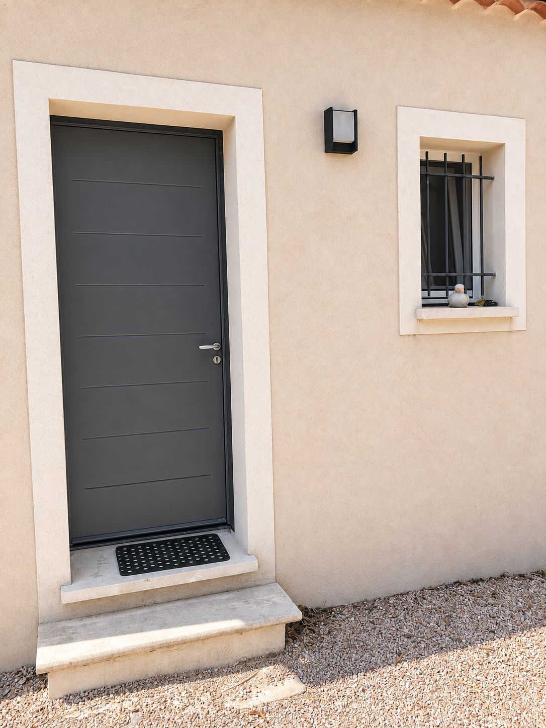 A modern building with a dark grey door, a small window with a metal grille, and a wall-mounted light fixture.