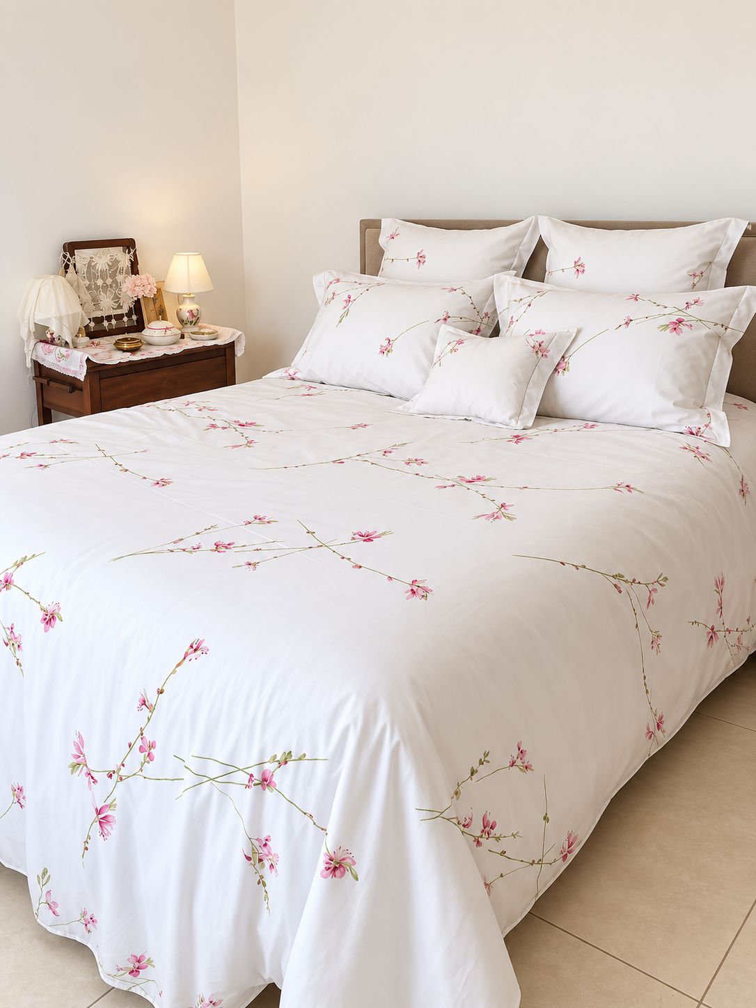 Un lit recouvert d’une couvre-lit à motifs floraux, agrémenté d’oreillers assortis, une table de chevet avec une lampe et un miroir orné d’un rideau en dentelle.