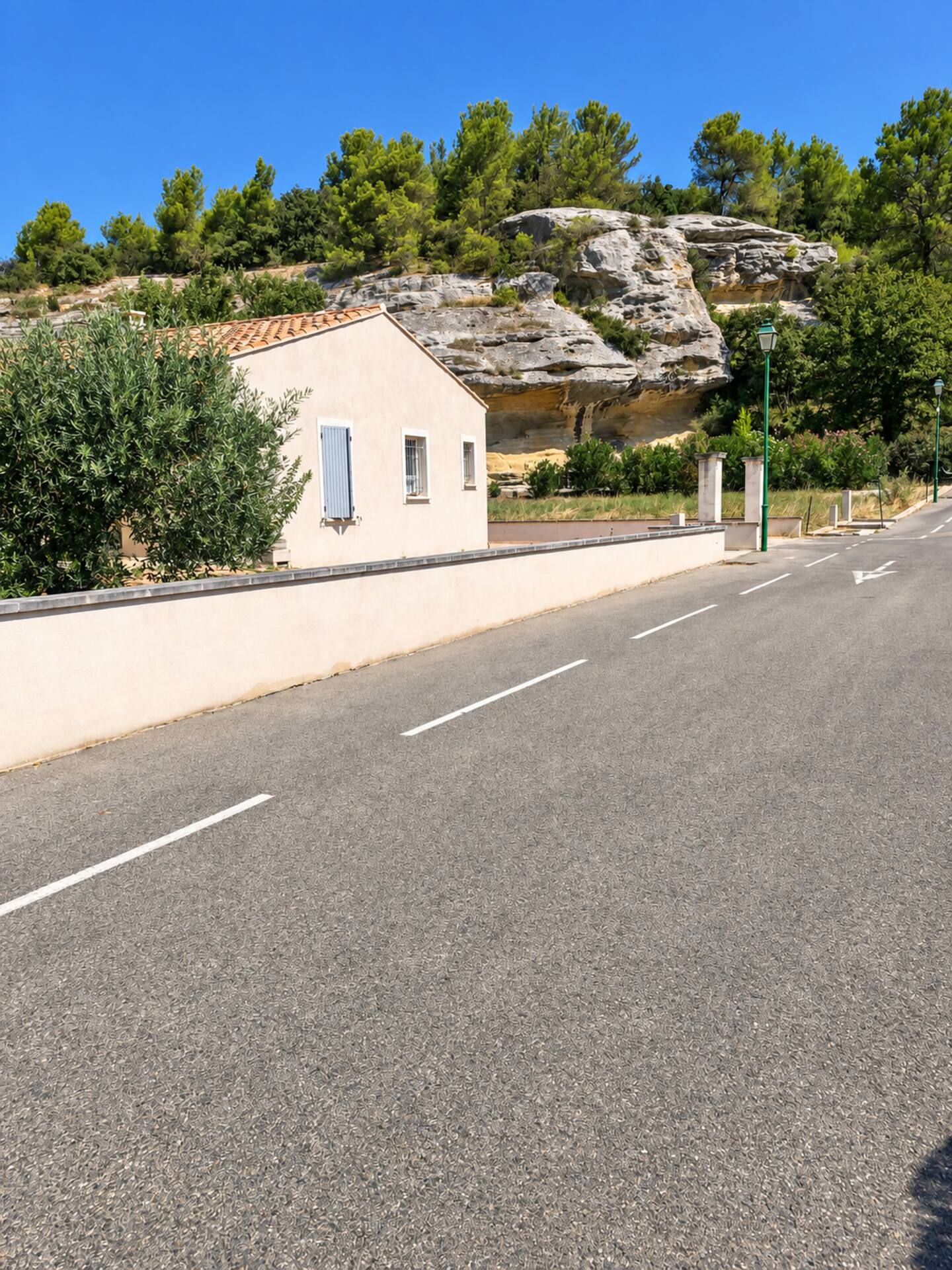 A road with a white building on the left, a rocky hill in the background, and trees.