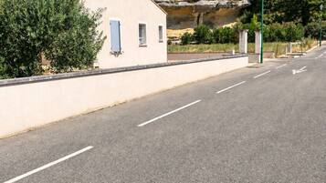 A road with a white building on the left, a rocky hill in the background, and trees.