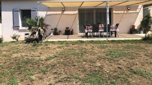 Une maison dotée d’un patio comprenant une table et des chaises, entouré de plantes en pot et agrémenté d’un espace salon abrité.